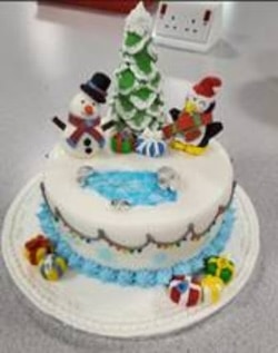 An iced snowman and penguin sit atop a cake, alongside a Christmas tree, beside an ice fishing hole.