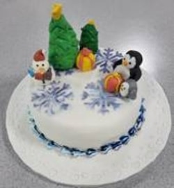 An iced snowman and penguin sit atop a cake, alongside two Christmas trees and some large snowflakes.